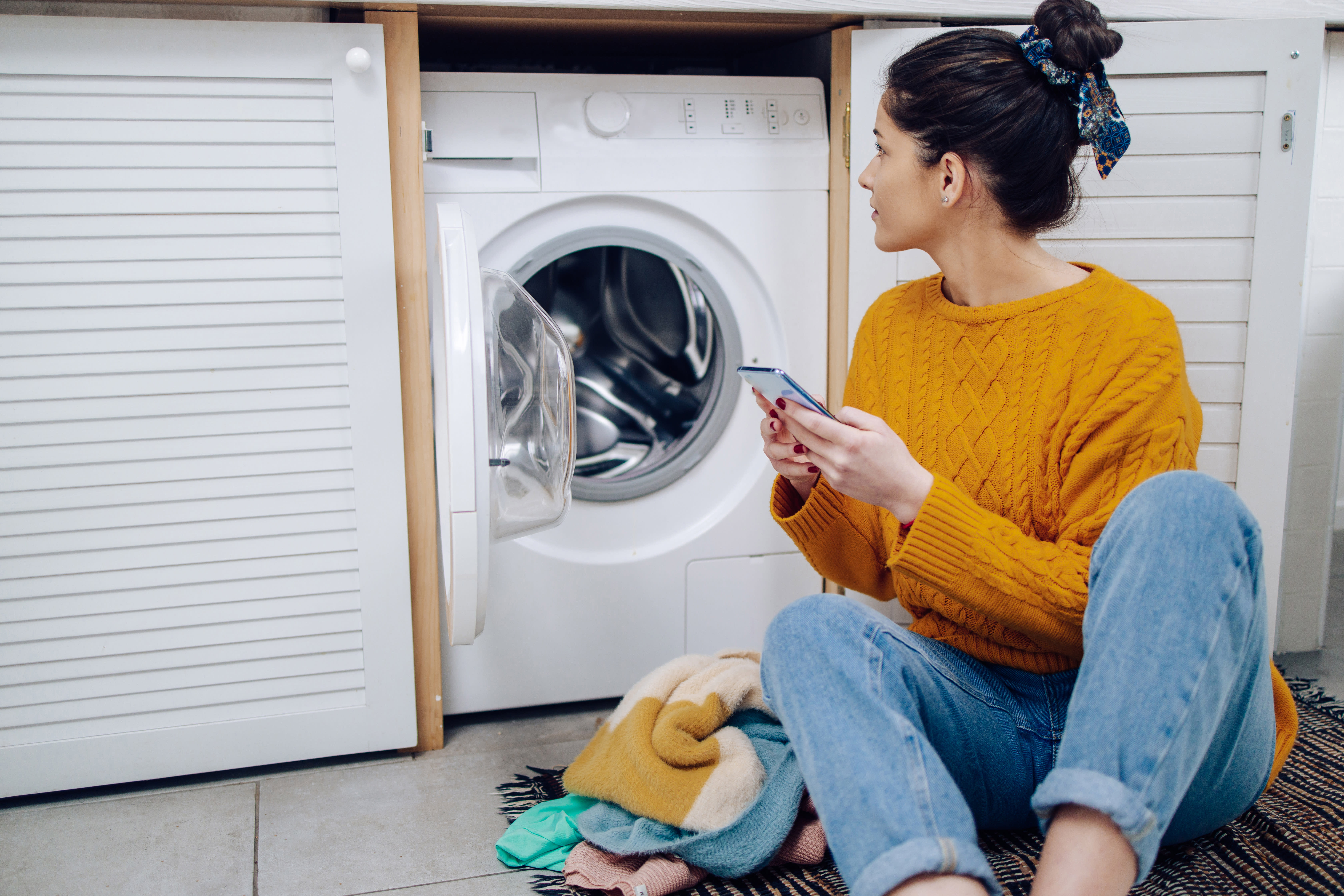 Young woman using a cell phone to adjust her smart washing machine in a laundry room