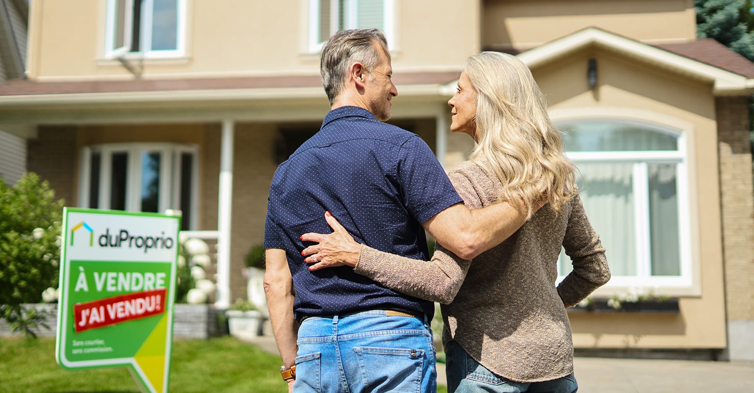 Un couple souriant à l'extérieur de leur maison, se regardant avec un panneau vert 'J'ai vendu' de DuProprio, illustrant leur succès dans la vente, tout sur la vente