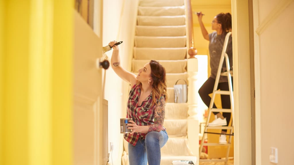 Two young women are painting a wall in yellow. One is kneeling, the other is on a stepladder. There is a staircase.