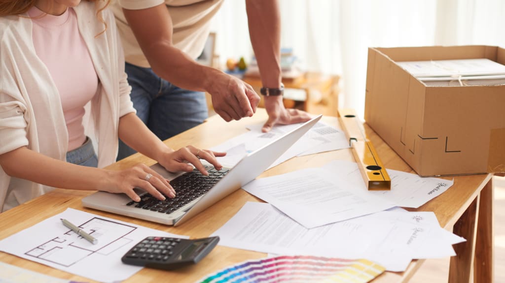 Two people consulting a laptop on a desk with building plans and colour charts