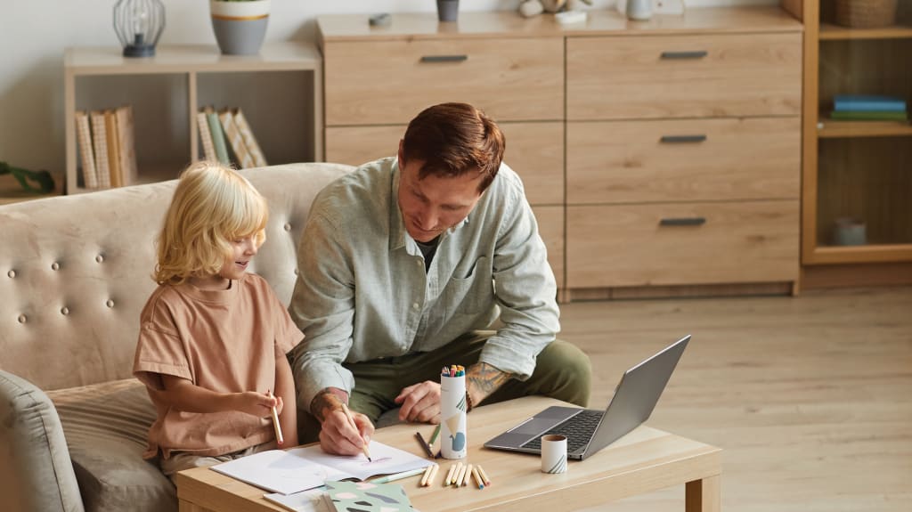 Un père et son enfant dessinent ensemble dans un salon de style scandinave, avec ordinateur portable et crayons à colorier sur la table