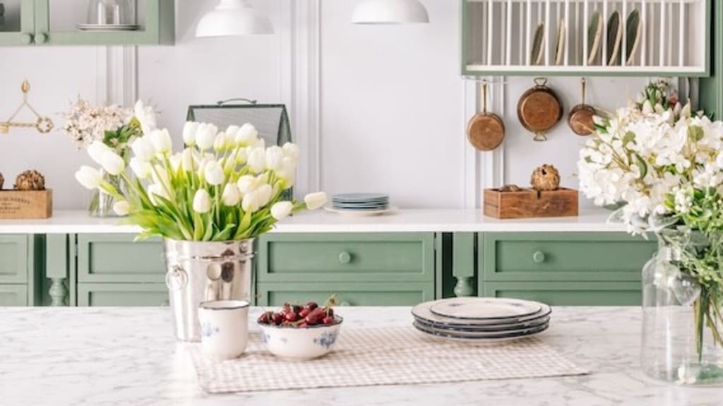 A kitchen painted sage and white with a marble central island and open cabinets