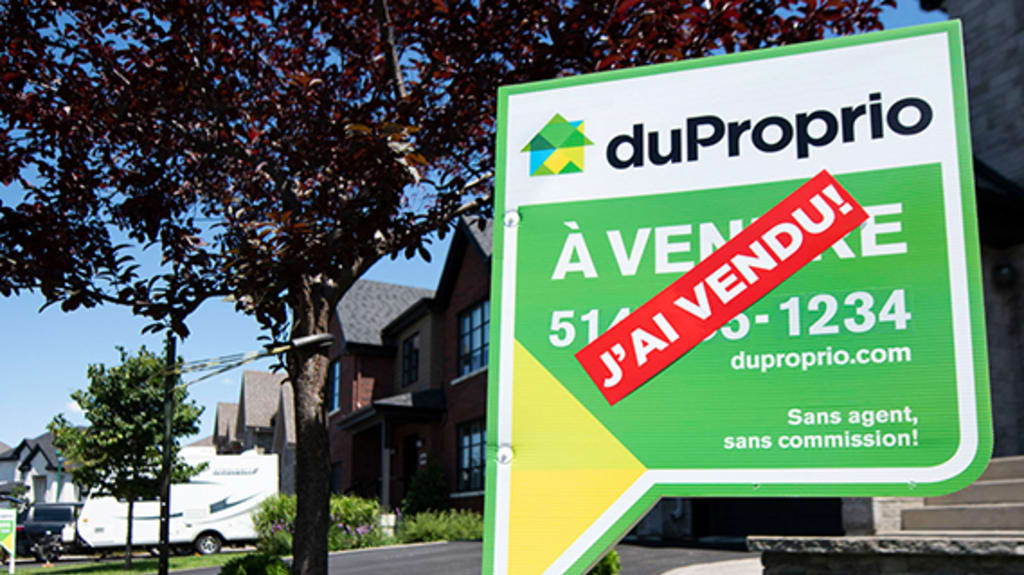 DuProprio sign with the "Sold!" sticker on the lawn of a dark gray stone house