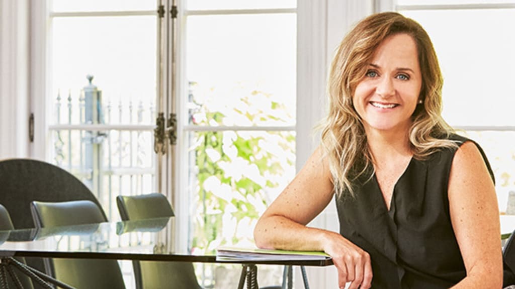 Brigitte Lafleur assise sur une chaise noire dans une salle à manger lumineuse comprenant de grandes fenêtres qui s'ouvrent sur une terrasse