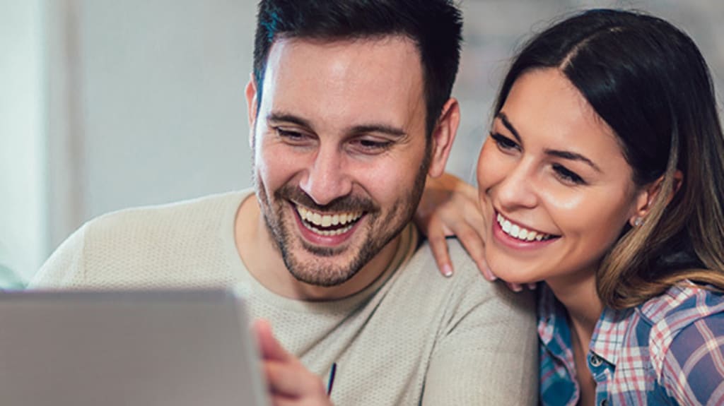 Un homme et une femme qui regardent un écran d'ordinateur en souriant et qui s'informent sur le Régime d'accession à la propriété