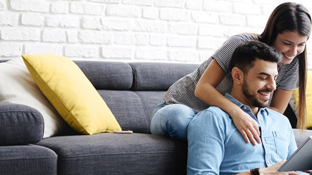 Couple dans un salon au mur de briques blanches installé sur un divan gris et regardant un document pour se préparer à faire une offre d'achat