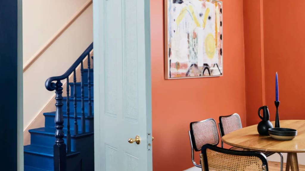 Salle à manger table bois, chaise en vannerie, murs orange, escalier bleu