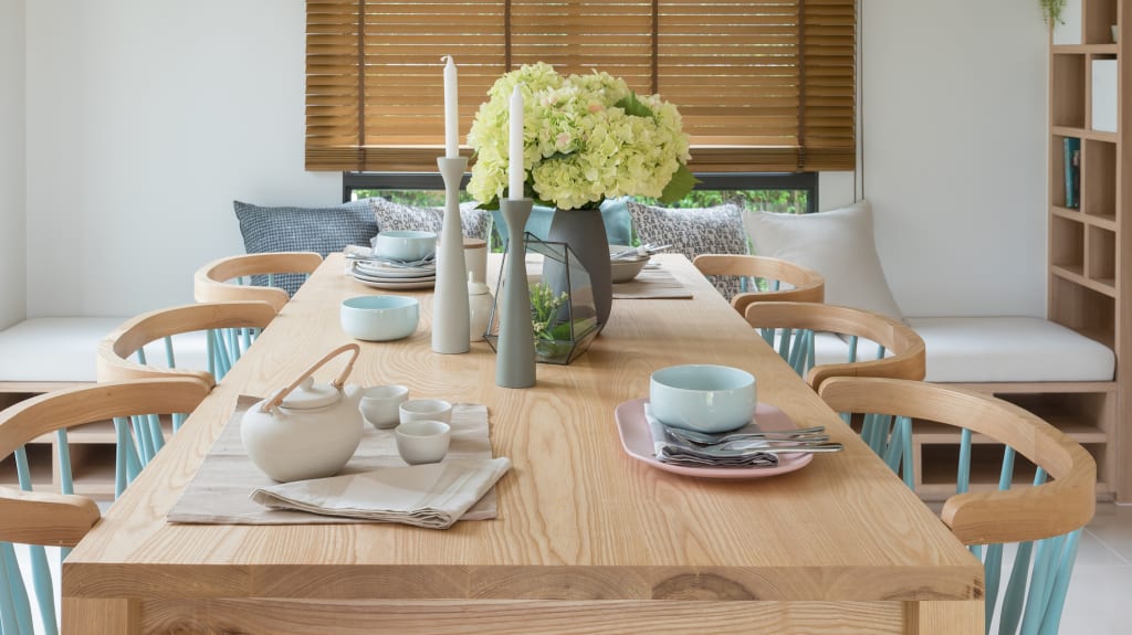 Dining room table in pale wood, six chairs with pale blue bars, fine porcelain tea service and vase with hydrangeas.Rustic wood dining table with four mismatched chairs. White plates. Several candle holders. Plants on table