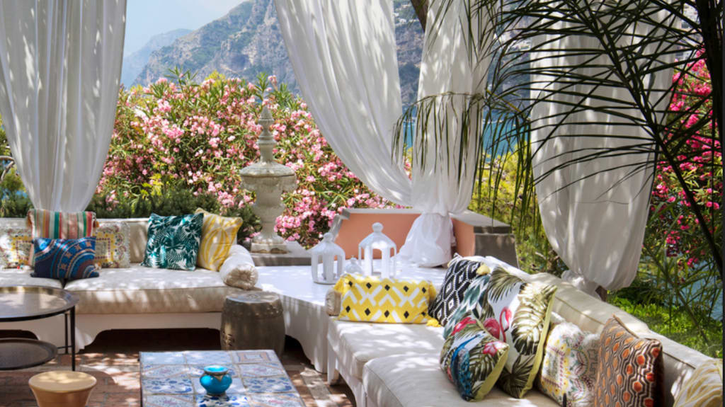 Déco voyage terrasse fleurie avec canapés couverts de coussins imprimés multicolores, montagnes en arrière-plan