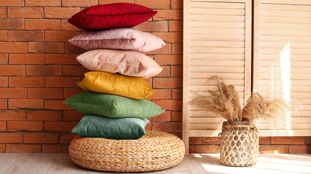 Pile de coussins de couleurs tendances variées sur pouf, vase avec de l'herbe de pampa posé sur le sol, devant un mur de briques