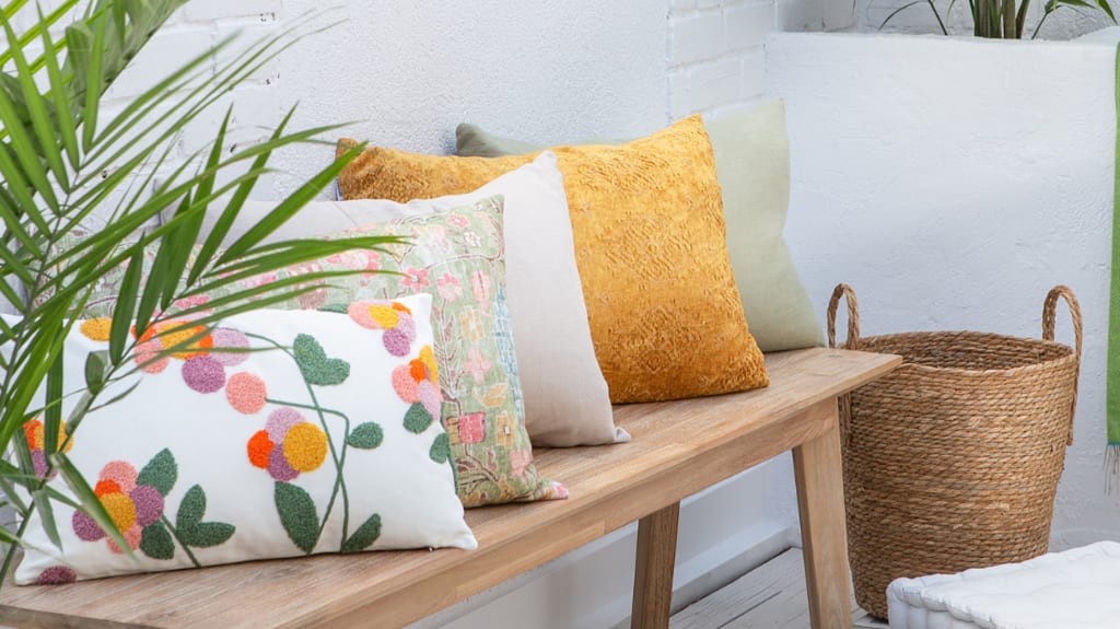 Yellow, beige and floral cushions on an outdoor bench, a wicker basket on the ground and a green blanket on a low wall