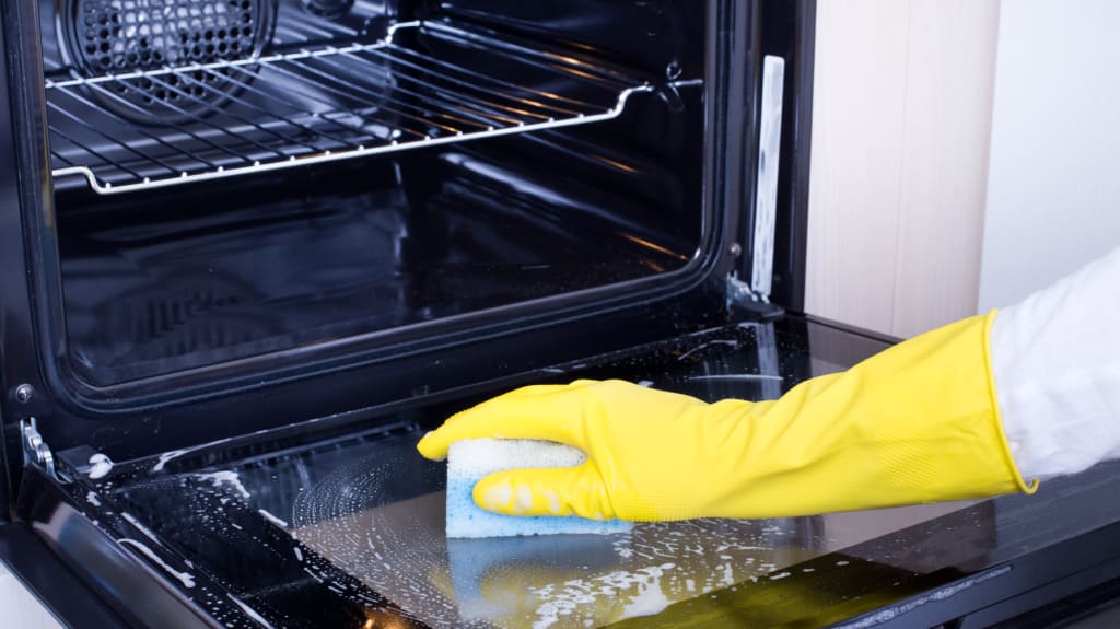 Personne avec gant de vaisselle jaune qui lave l'intérieur de son four à l'aide d'une éponge et d'un mélange savonneux.