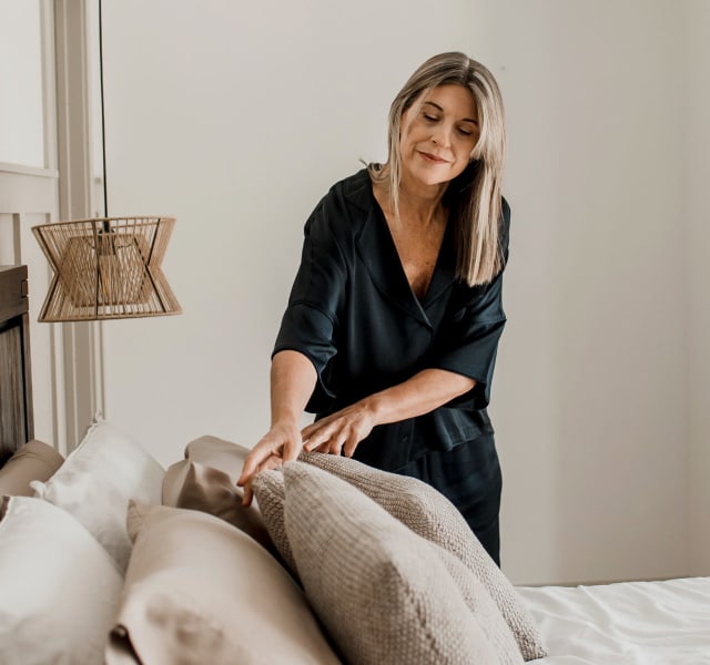 A blonde woman puffing some pillows on a bed