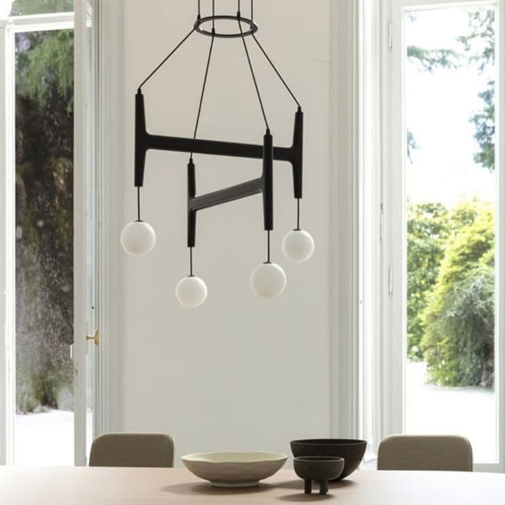 Salle à manger avec grandes fenêtres ouvertes sur jardin. Table en bois, vases et lampe suspendue avec globes en verre blanc.