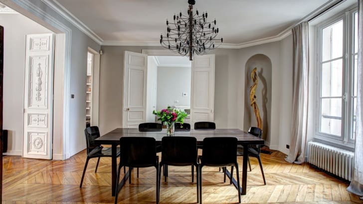 Elegant dining room, doors with moldings, square table and eight black chairs. Black light fixture with multiple branches. Abstract sculpture in an alcove.