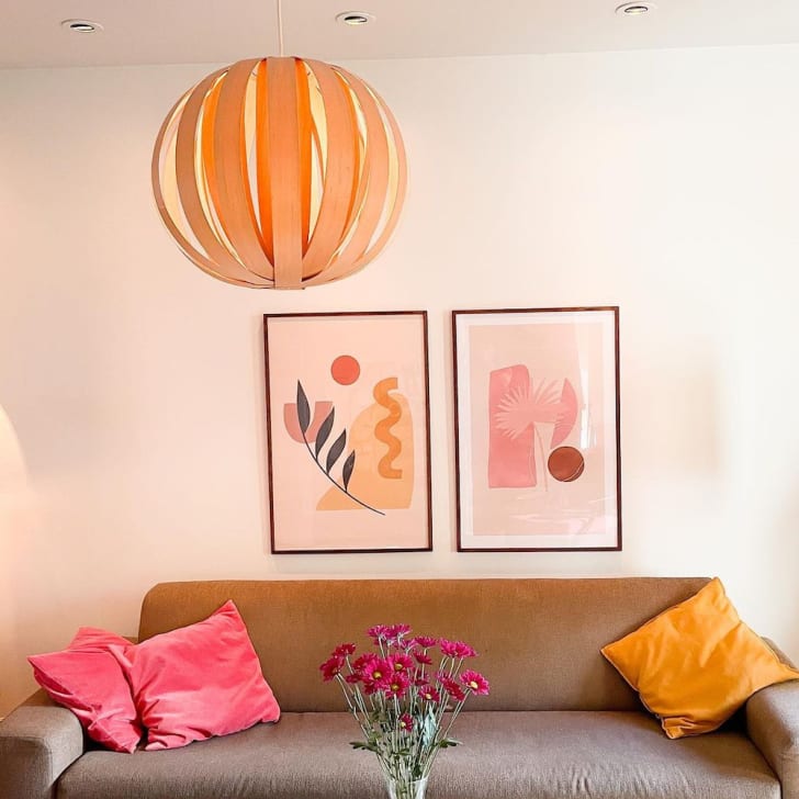 Salon, canapé brun avec coussins roses et orangés, affiches encadrées, lampe suspendue en lattes de bois de forme sphérique