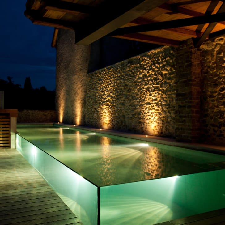 Piscine étroite trois parois de verre, éclairage de nuit, mur de pierre et terrasse en bois