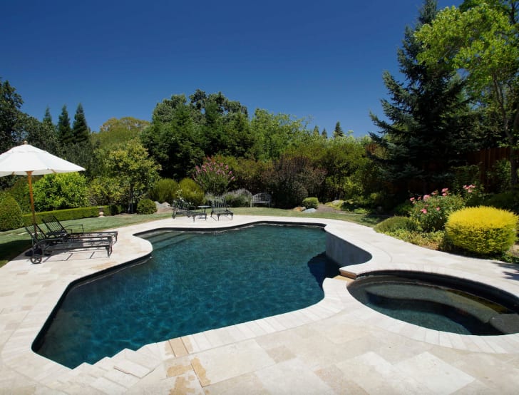 Belles piscines bassin forme libre avec spa ovale à côté, fond bleu foncé, parasol et deux chaises longues, arbres et arbustes soignés