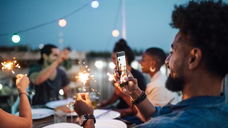 Jeune homme prenant une photo de cierge magique avec des amis assis à table pendant une fête en soirée