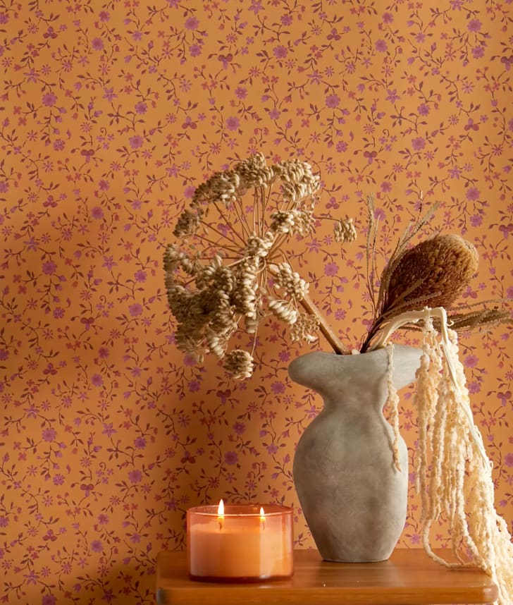 Déco voyage fleurs séchées dans un vase en grès aux formes incurvées, bougie à deux mèches allumées, mur couvert de papier peint orange abricot avec fleurs violet