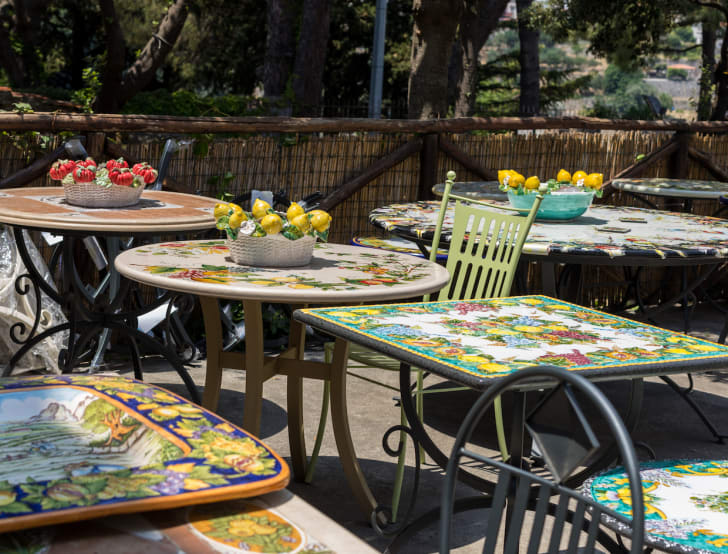 Déco voyage, terrasse avec plusieurs tables à plateau en céramique orné de motifs de fruits, plats en céramique ornés de citrons et de tomates, chaises en fer forgé, clôture en bambou
