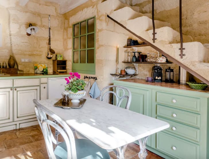 Déco voyage cuisine traditionnelle du sud de la France, murs en pierre, bahut vert pâle, table en marbre et fer forgé, chaises bistro blanches et bleues, vaisselle sur étagères
