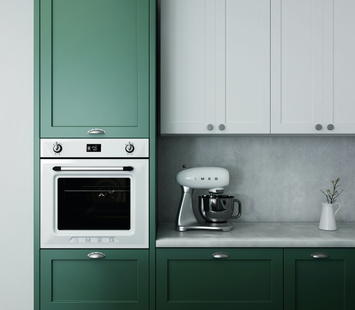 Melamine kitchen cabinets painted green and white, small built-in oven, Smeg mixer and pot of dried flowers on countertop
