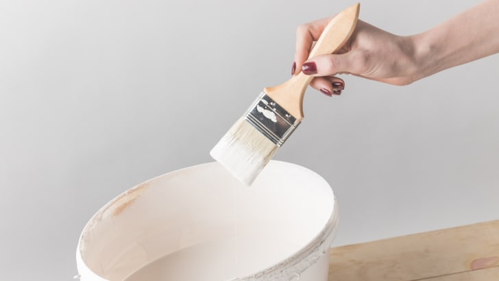 Cropped image of a person holding brush in white melamine paint above bucket