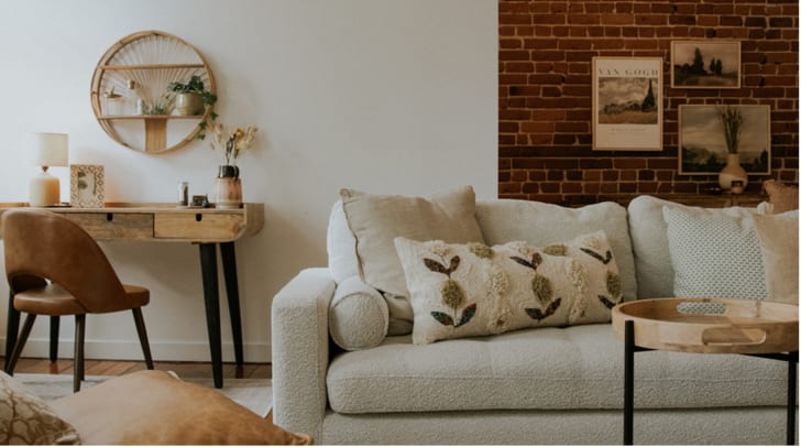 Vue sur un salon incluant un divan gris devant un mur de briques, coin bureau à gauche avec chaises et items décoratifs pour la maison