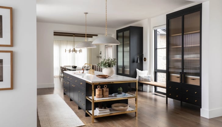 Vue sur l'ilot d'une cuisine avec une robinetterie dorée, luminaires blancs suspendus, armoires noires vitrées, accessoires décoratifs