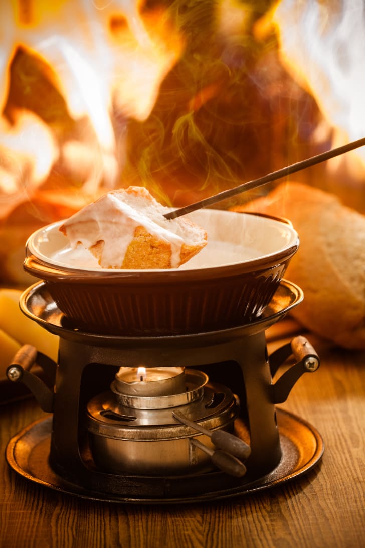 Cheese fondue pan including a burner, fondue stick with a piece of soaked bread and flames in the background