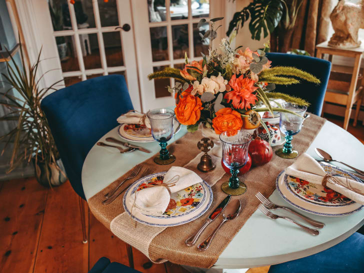 Bouquet de fleurs automnal déposé sur une table à manger ronde, chemin de table en lin, coupes translucides bleues, ustensiles et vaisselle fleurie