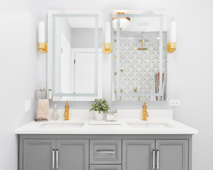 Bright bathroom with two mirrors, gray vanity, gold faucets and fixtures, granite countertop