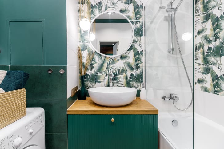 Petite salle de bain moderne dans les teintes de vert et de blanc dont le mur est décoré d’un adhésif mural aux motifs de feuillage, incluant une sécheuse, un meuble lavabo et une baignoire-douche