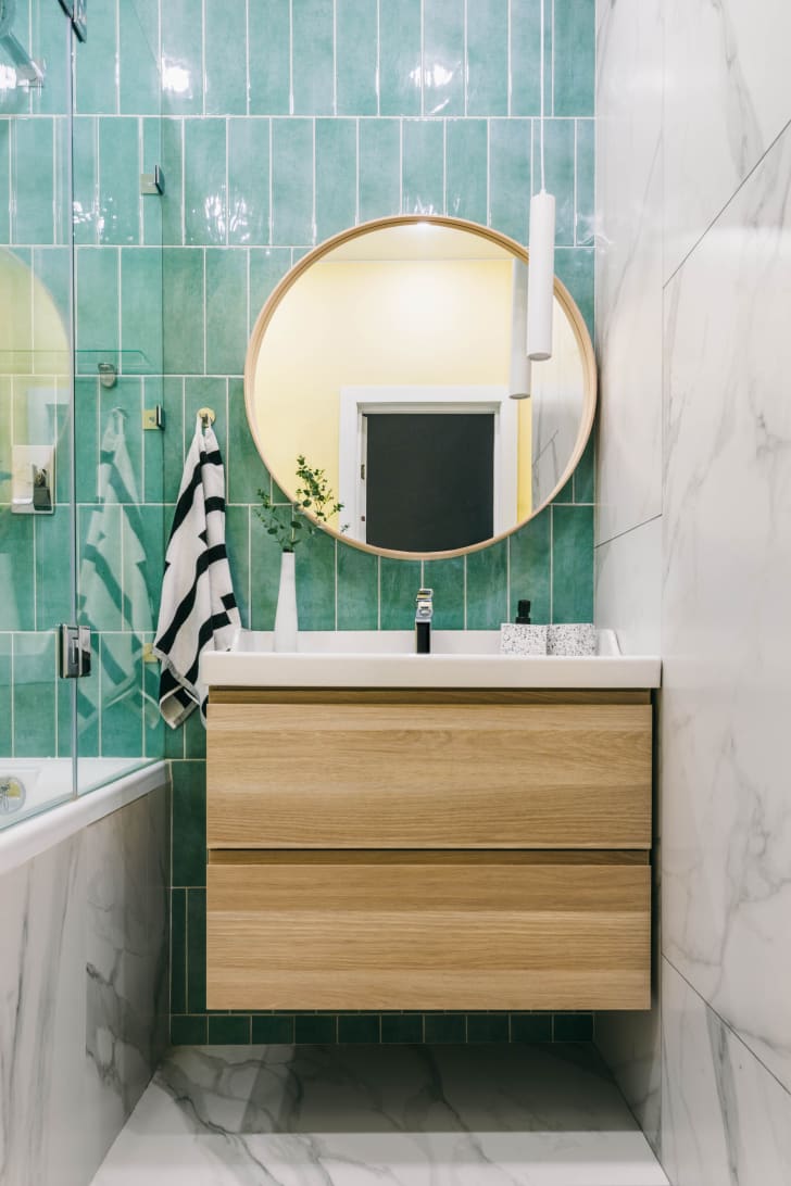 Round mirror above a floating wooden vanity in a small bathroom with a blue-green tiled wall and marble floor 