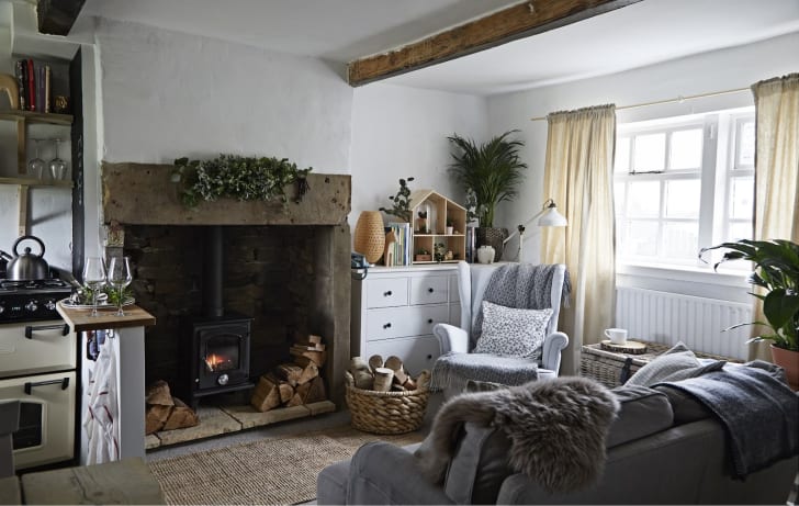 Décoration de salon cottagecore, avec foyer, poutres au plafond, et grosses couvertures de laine posées sur les fauteuils gris