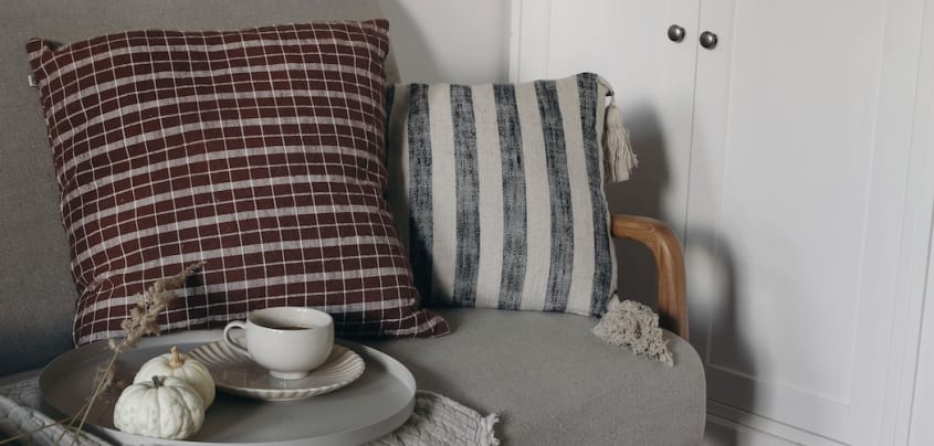 Canapé gris décoré d’un coussin à carreaux et d’un autre ligné, plateau, petites citrouilles et tasse de thé