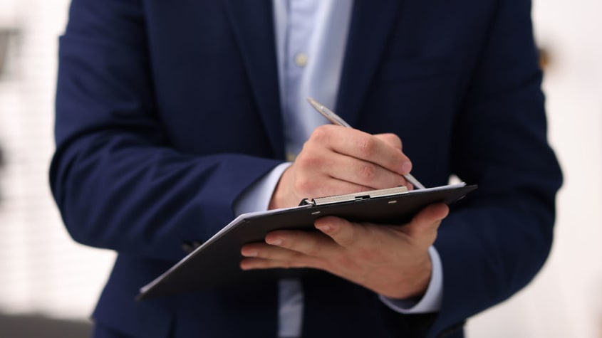 Person wearing a dark blazer writing on a form placed on a clipboard